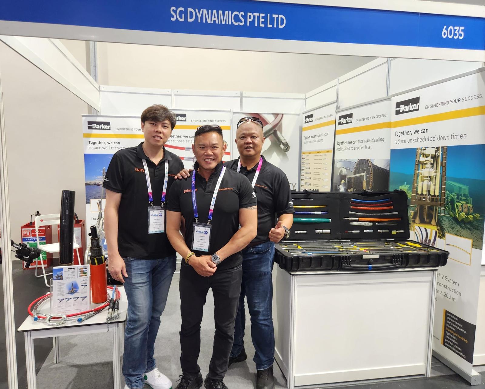 Three team members from SG Dynamics Pte Ltd stand at their exhibition booth, showcasing equipment and wearing name tags.