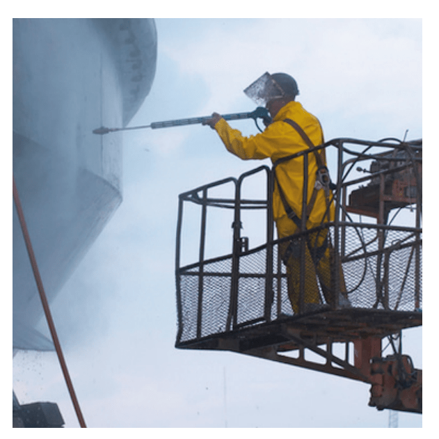 Worker in yellow gear using a high-pressure water jet for industrial cleaning on large metal equipment or ship surface.