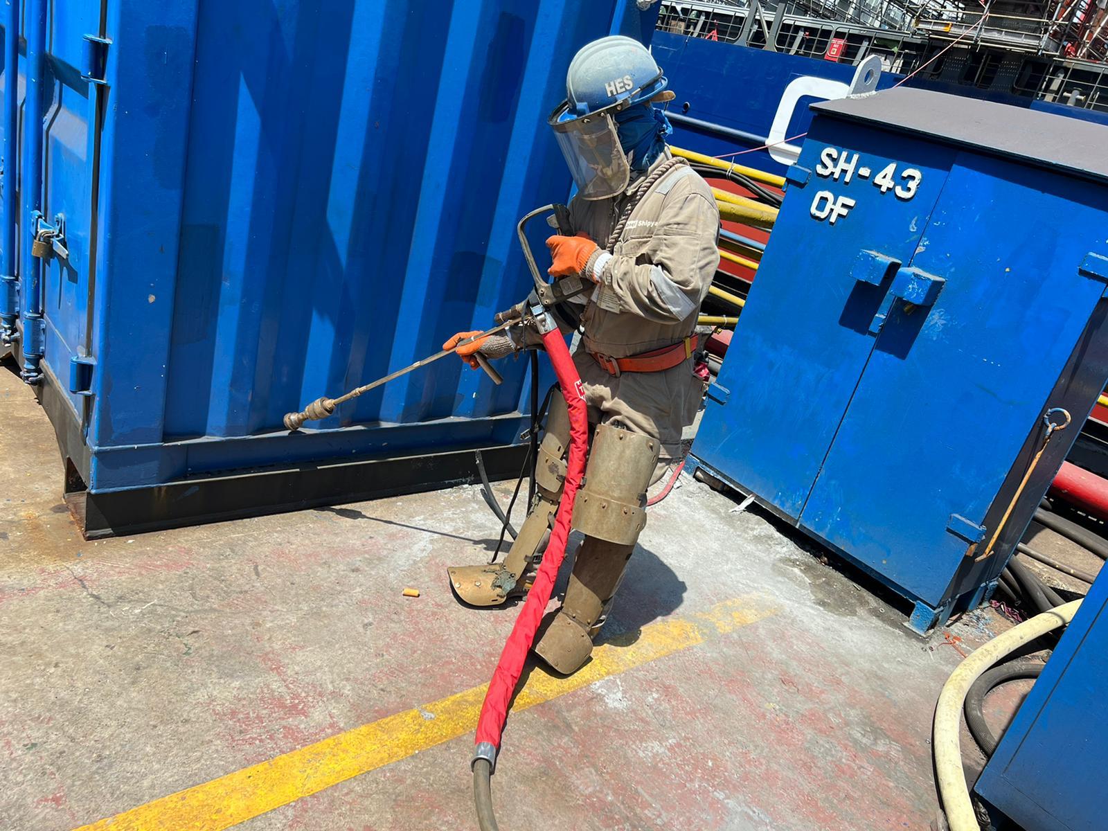 Worker in protective gear using a red hose near a blue container, likely for cleaning or maintenance tasks.